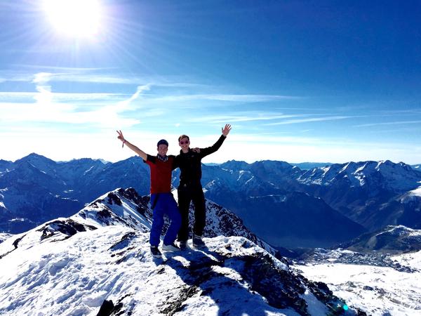Två killar håller varandra om axeln på en bergstopp med strålande sol i bakgrunden.