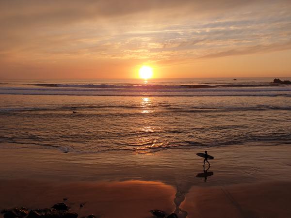 En ensam surfare går längst stranden och solen går ner över horisonten.