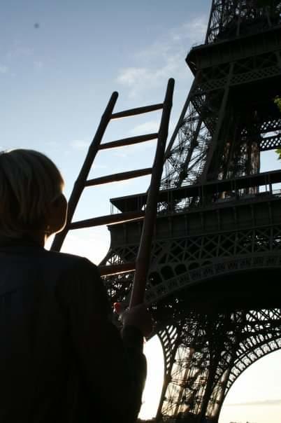 En kvinna står vid foten av Eiffeltornet, och i händerna håller hon en stege riktad mot tornets topp.